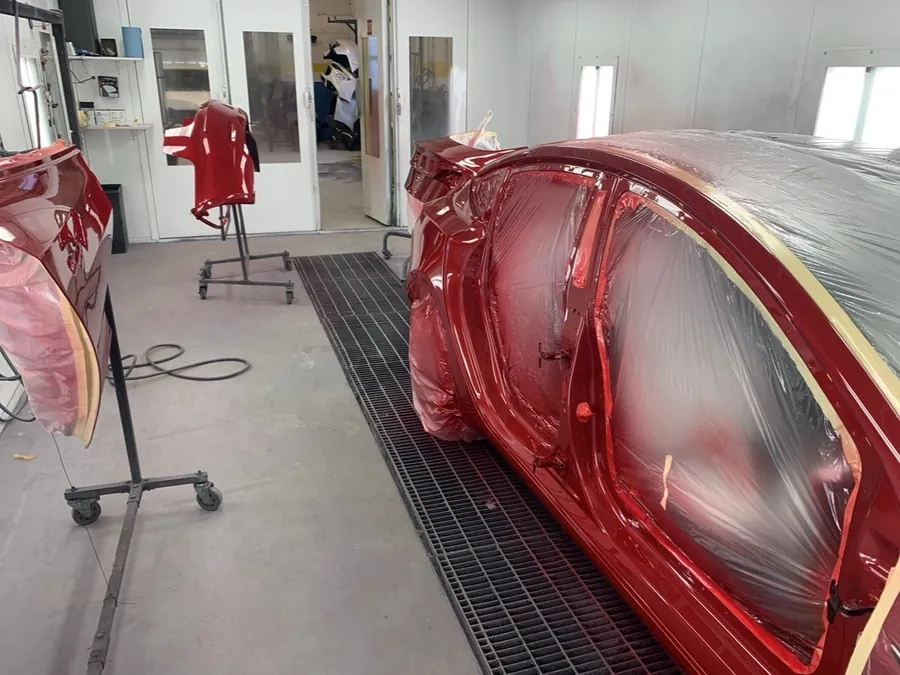Red car in the paint booth with its bumper and door panels removed and refinished separately on stands